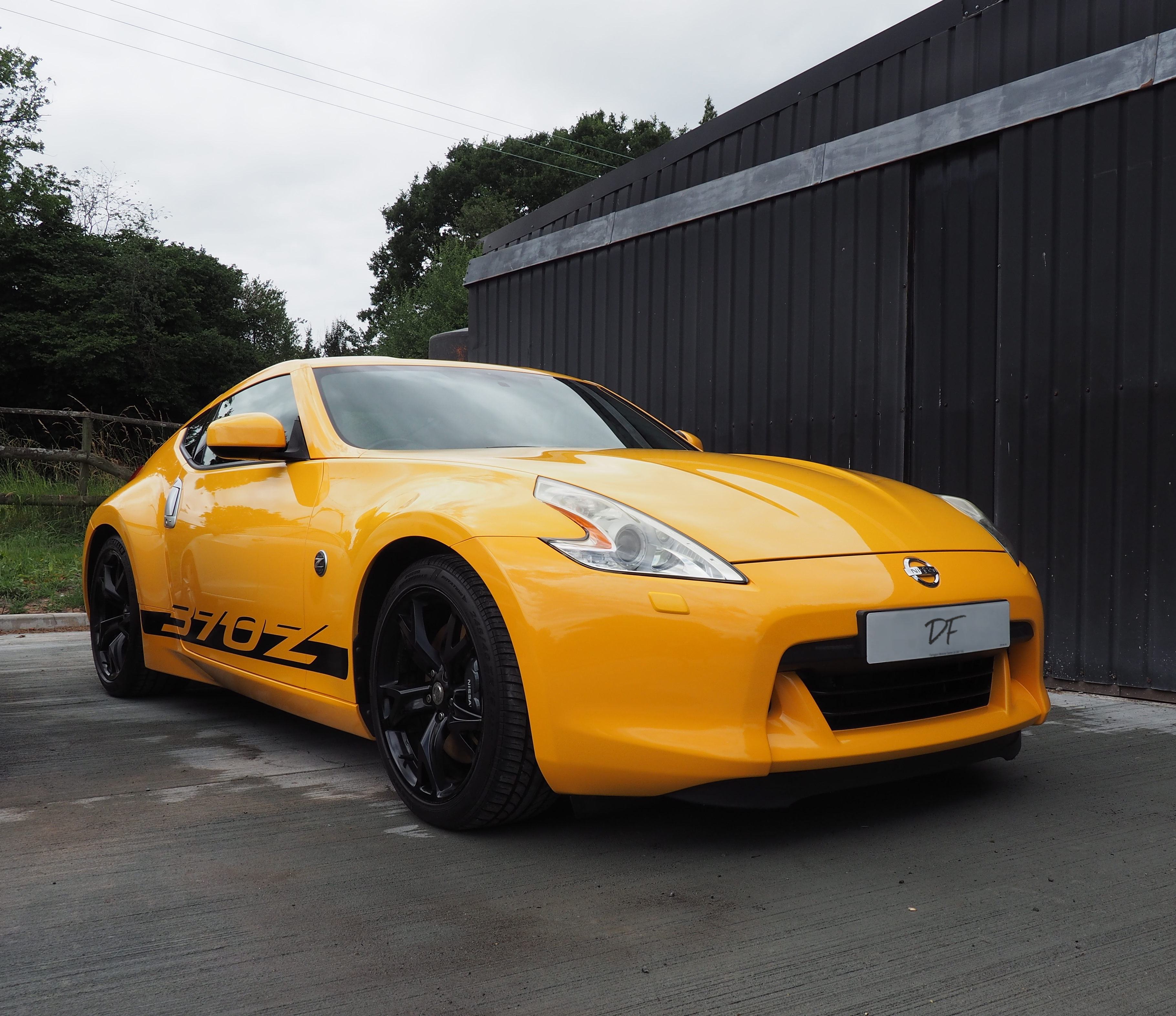 A rare ultimate yellow 370z, in-front of a contrasting dark grey industrial sliding door, the sky and surroundings reflecting of its pristine paintwork, enhanced by a generous layering of paint sealant and graphene based wax.